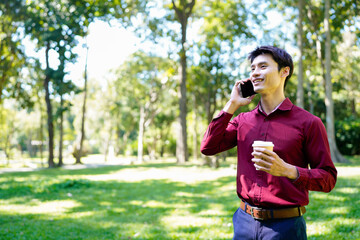 Fototapeta premium Young asian man with takeout coffee communicating on smartphone at urban park, blank space