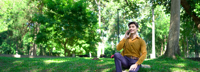 Happy asian man using a smartphone in a park, enjoying a pleasant conversation on a sunny day, surrounded by lush greenery and modern architecture.