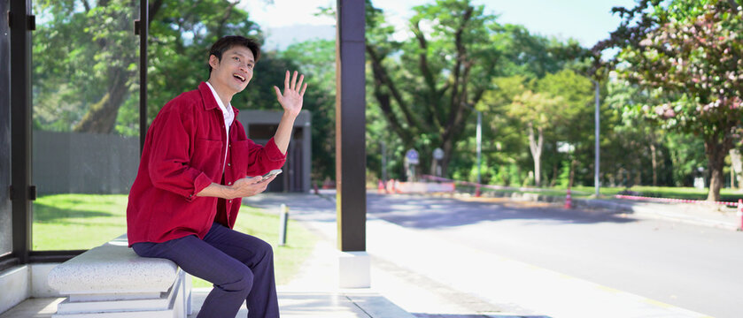 Young man with smartphone waiting for public transport at bus stop - Powered by Adobe