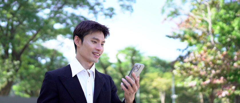 Young asian businessman in black suit with takeout coffee using smartphone at urban park, blank space - Powered by Adobe
