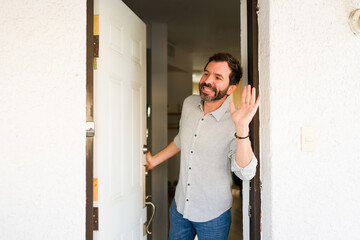 Happy man welcoming guests or saying goodbye, standing in doorway of home, smiling and waving hand