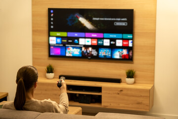 Woman uses a remote control to control a modern, large LED TV. Entertainment streaming service that allows users to enjoy wide selection of award-winning movies, TV series, and programs.