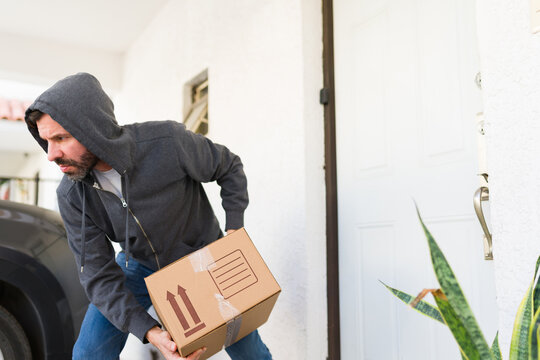 Criminal man in a hoodie taking a cardboard box from a residential porch, illustrating package theft and home security issues