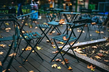 Dark metal bistro chairs and tables sit on a wet wooden deck, scattered with orange and yellow fallen leaves on a cool autumn day.