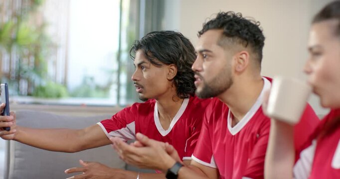 Friends in red jerseys getting score update on phone gesturing sipping mug on light-colored sofa