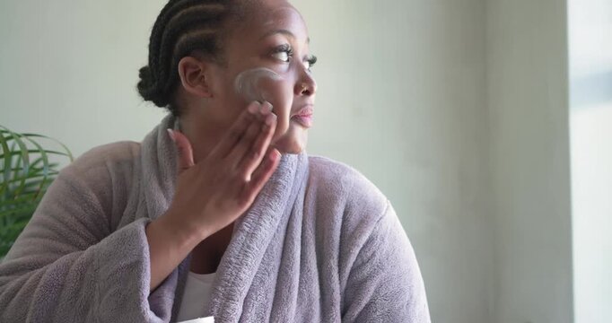 Window light guiding African American woman at home opening jar and dabbing cream hydrating skin
