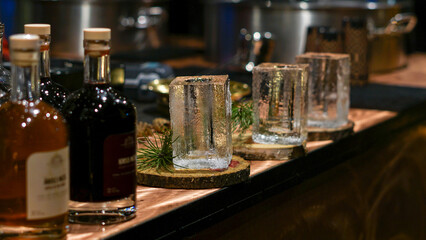 A rustic bar scene featuring whiskey bottles beside drinking glasses made from blocks of ice on a wooden counter. Christmas market decorations, festive shopping.