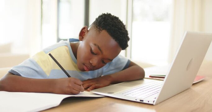 Resting arm on table, African American boy scribbling with pencil and viewing laptop info by window