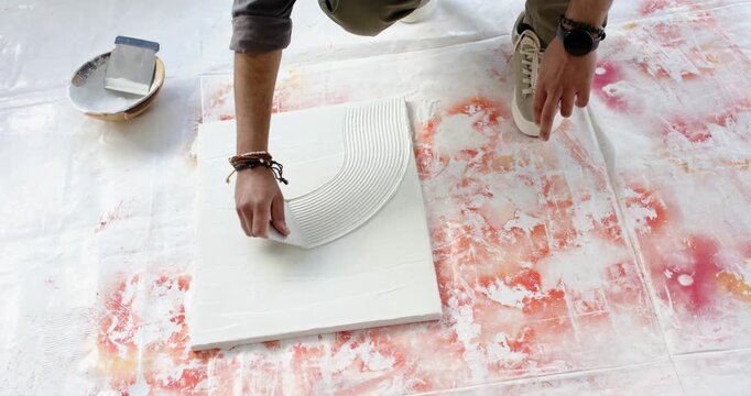 Arriving beside canvas, man is scooping compound with notched trowel and shaping ridges in workshop