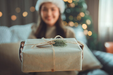 Woman giving Christmas gift wrapped in craft paper with natural decoration, festive holiday atmosphere with warm lights and winter celebration mood in cozy home interior