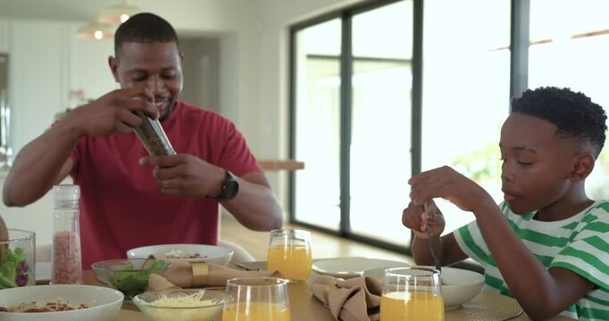 African American father seasoning pasta with mill as son pausing while mother serving salad at home