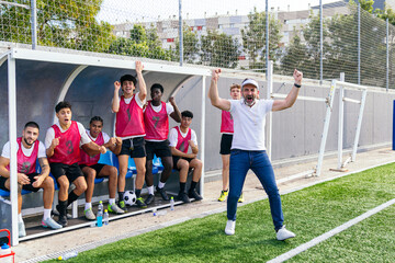 Male coach and young football players celebrating success on the bench during a match, showing excitement and sportsmanship