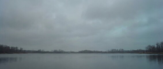 Moody winter lake landscape under vast gray overcast sky reflecting bare trees solitude