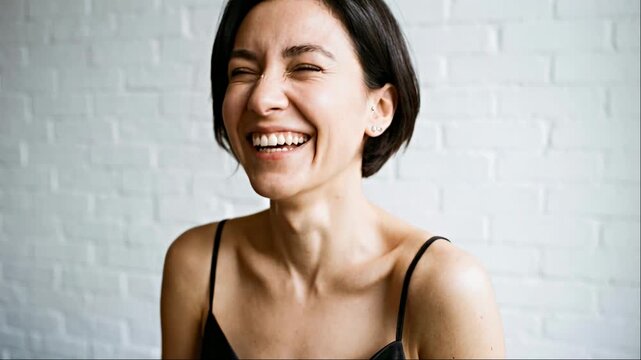 Joyful young woman with short dark hair laughing wearing a black silk camisole against white brick wall