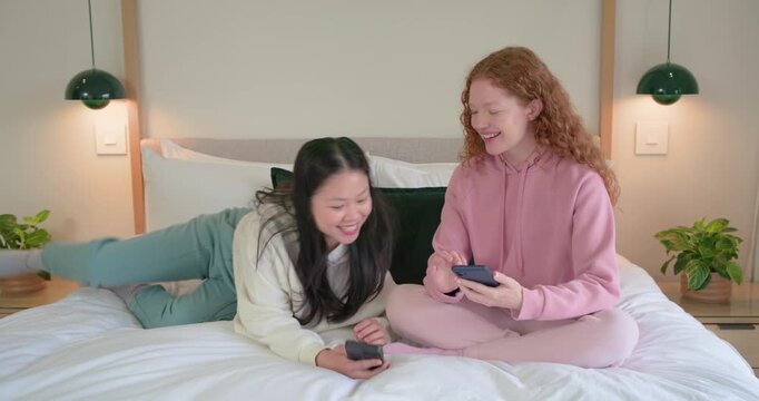 Diverse female friends on bed tapping phones, second woman entering, showing content, laughing