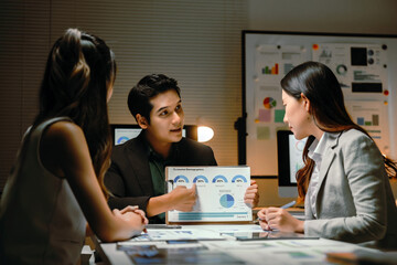 Business colleagues working together, reviewing consumer demographics data and charts during a meeting in a dimly lit office