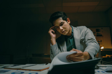 Young professional man analyzing documents, feeling stressed and overworked while staying late in dark office for a deadline