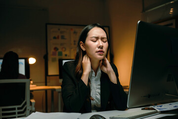 Young Asian businesswoman sitting at desk, eyes closed, massaging her aching neck, experiencing stress and burnout