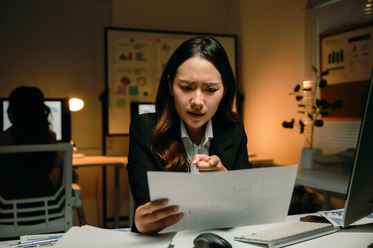 Asian businesswoman working overtime, feeling overwhelmed and frustrated while reviewing documents in a dimly lit office environment