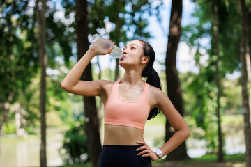 Young athletic woman drinking water from a bottle, refreshing after an intense workout or jogging session in a lush green park