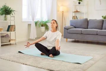 Fotobehang Lotusbloem Mature lady wearing sportswear sitting cross legged on sport mat in living room and meditating with closed eyes. Calm senior woman relaxing and practicing yoga breathing exercises in lotus position.  © Studio Romantic
