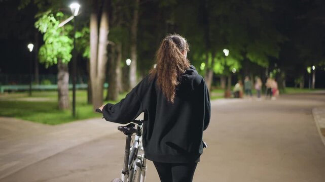 Woman Cycling Along Illuminated Treelined Avenue, Serene Night Journey Of Woman Pedaling Past Glowing Trees, Calm Evening Bicycle Ride Beneath Luminous Trees In Tranquil Urban Setting