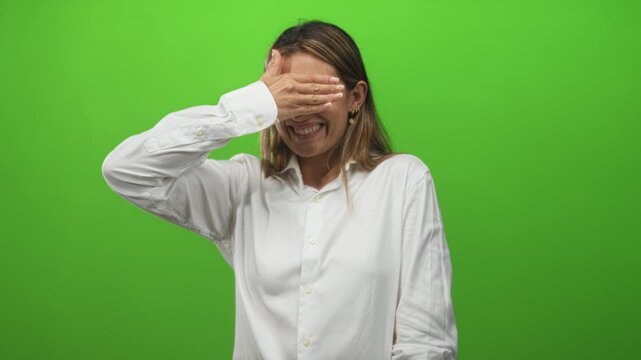 Young blonde woman wearing white shirt and small hoop earrings covers eyes with hand while smiling on green studio set; shy.