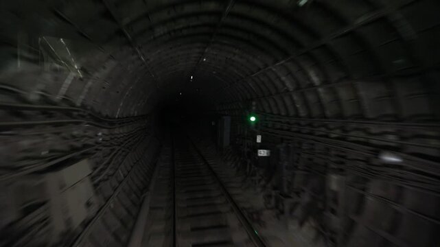 View from the rear of a moving subway train showing railway tracks. The train passes through a dark tunnel and arrives at a bright underground station platform.

