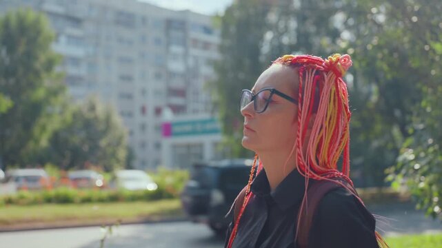 pensive caucasian woman with pink braids standing by apartment street, city buildings and trees in background, glasses, backpack, soft daylight, reflective mood, solitary urban portrait conveying