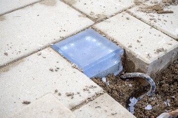 Transparent glass block embedded in paving stones with visible soil and wire. Landscape lighting, garden design, construction detail. Real photo