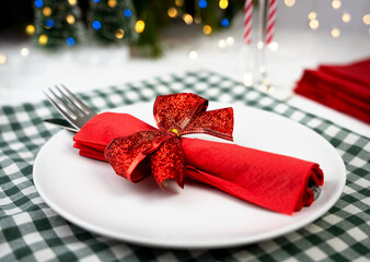 Elegant Christmas table setting with a red napkin wrapped in a glittery bow and silverware placed on a white plate, surrounded by festive lights and holiday decor. Close-up. Selective focus.