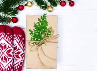 A craft paper gift decorated with green branches and twine, red knitted mittens on a white wooden background. Close-up. Copy space.