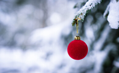 Red velvet Christmas ball hanging on a snow-covered evergreen branch outdoors. Soft winter background creates a peaceful and festive holiday mood.