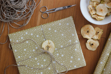 Christmas gift wrapped in gold paper and decorated with dried apple, dried apples in a bowl, ribbon and scissors on wooden floor. Christmas flat lay.