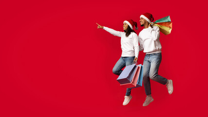 Two friends are joyfully jumping against a bright red background, wearing Santa hats and holding shopping bags, displaying their enthusiasm for the festive shopping season.