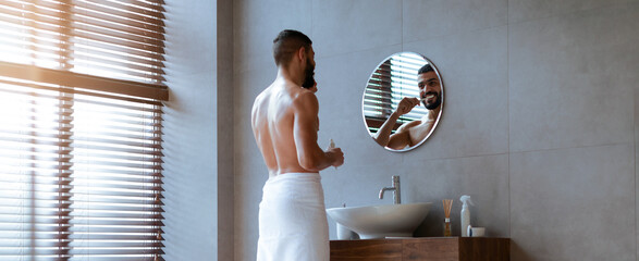 A man stands in a stylish bathroom wrapped in a towel, preparing for his day. He looks at his reflection in the mirror while grooming himself amidst modern decor and soft lighting.