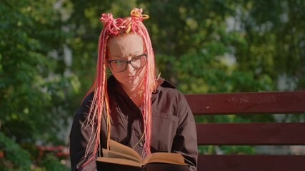 reader enjoying sunlight, individual with vibrant hair immersed in books outside, caucasian woman with bright pink plaits focused intently on her book amidst greenery on sunny afternoon