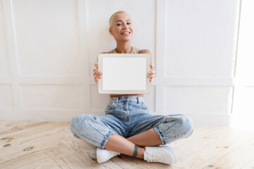 Excited woman showing modern digital tablet with empty screen, newest pad with blank display, mockup. Lady sitting on floor over light wall showing various mobile apps for tablets