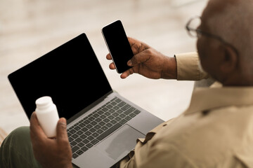 A senior man sits at home, focusing on his smartphone while holding a bottle of medication in one hand. His laptop rests open on a table in front of him.