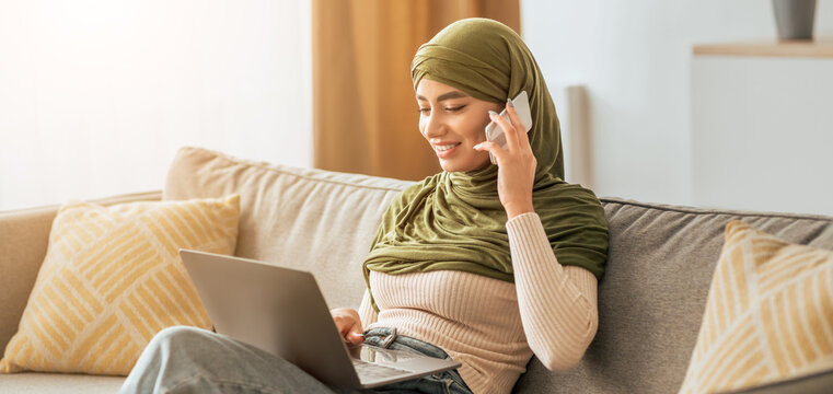 A young woman in a green headscarf sits comfortably on a beige sofa. She is smiling while using her laptop and talking on the phone, enjoying her work at home in a bright atmosphere. - Powered by Adobe