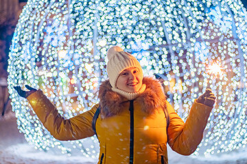 Young woman in knitted hat holds sparkler in her hands. Night festive street, shiny city decorations for Christmas.