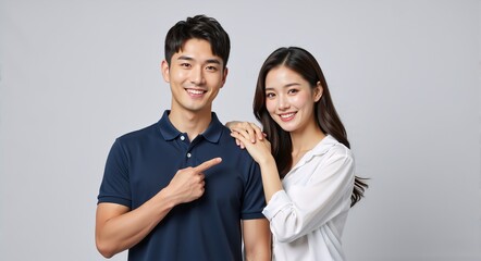 Happy young Asian couple standing together with man pointing finger at woman. Cheerful boyfriend and girlfriend portrait isolated on grey background. Recommendation concept
