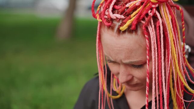 tearful woman with vibrant dreadlocks outdoors, expressive woman with colorful locks shows emotion outside, crying woman with bright dreadlocks covering face amidst park setting with tears