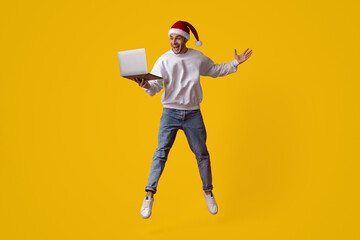 A joyful individual is leaping in the air while wearing a festive Santa hat and a cozy sweater. They hold a laptop in one hand, showcasing their enthusiasm.