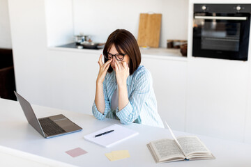 A woman sits at a kitchen table, wearing glasses, feeling stressed while working on her laptop. Notes and a cookbook are beside her. She looks overwhelmed during her task.