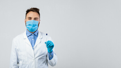 Millennail male doctor in protective mask and gloves holding sterile swab stick for coronavirus PCR test on grey studio background. Young medic with tube for nasal and oral covid diagnostic procedure