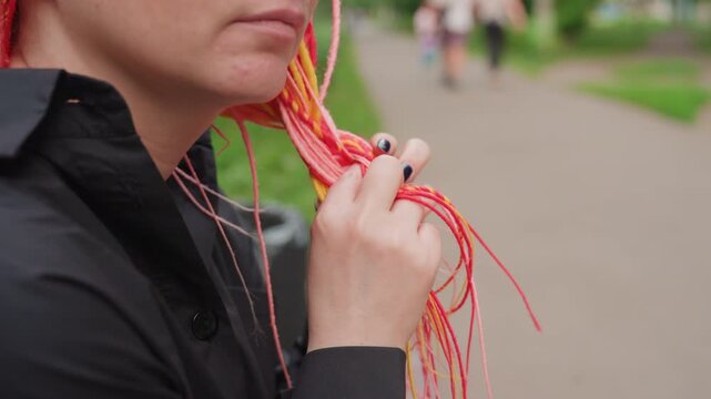 woman holding colorful earphones in park, pensive expression and fidgeting with braided cords, neon manicure visible, black coat, blurred pedestrians in background, intimate urban solitude and quiet
