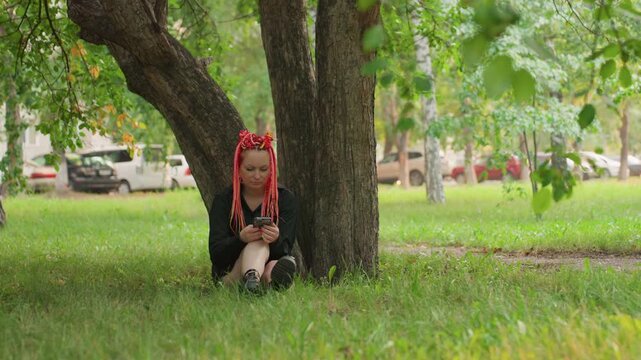 caucasian teen sitting under tree looking at phone, red braids, black outfit, grass foreground, distant cars, quiet afternoon, solemn expression, scrolling messages, reflective mood, urban park