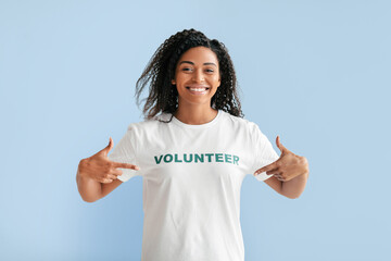 A cheerful woman wears a white t-shirt with the word VOLUNTEER printed in green. She smiles broadly and points at her shirt, showcasing her enthusiasm for community involvement.