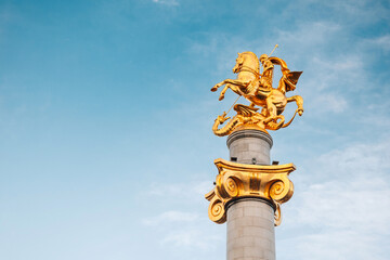 Naklejka premium 1 November 2025, Tbilisi, Georgia: Golden statue of Saint George on a tall column in the city center, representing victory and freedom